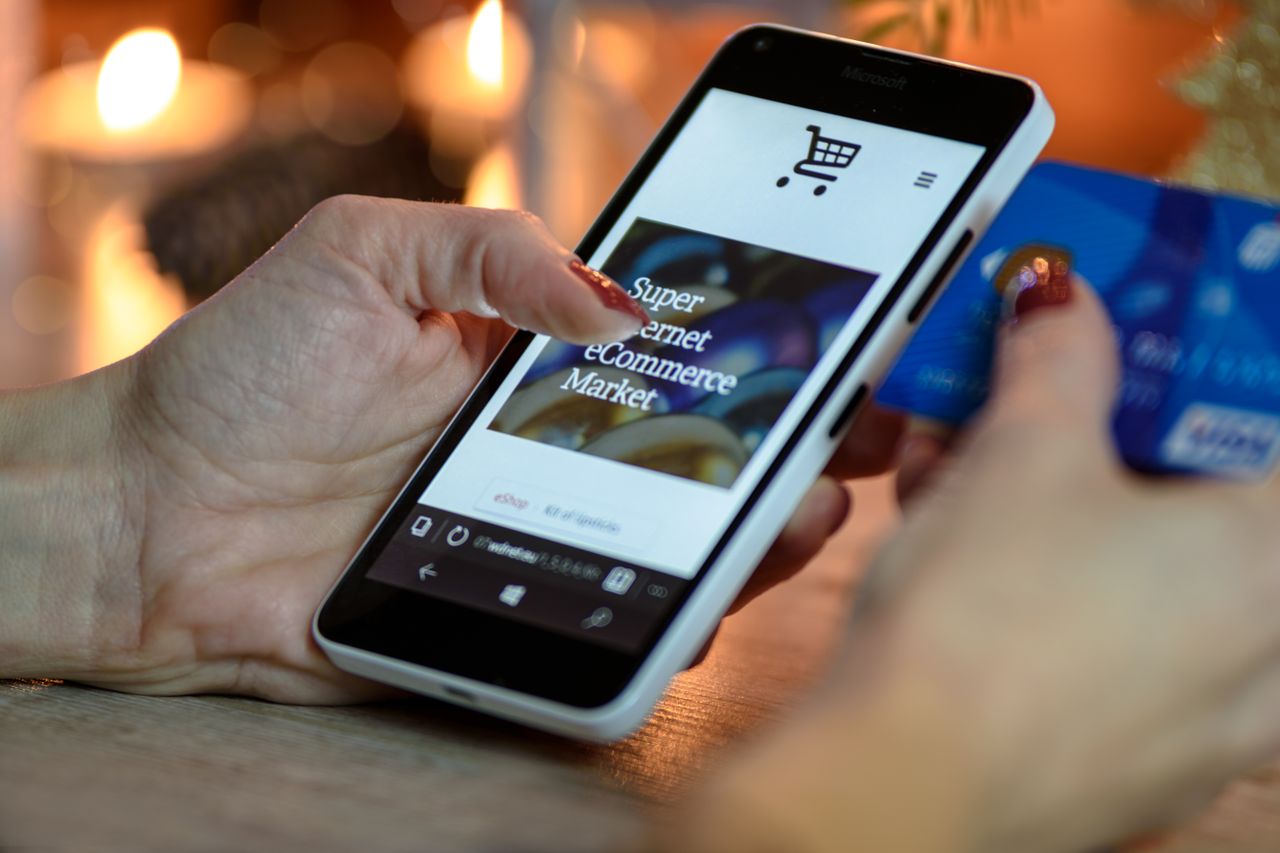 Woman using smartphone for online shopping while holding a credit card, with festive background lighting.